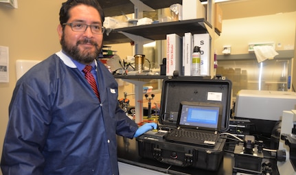 Dr. Luis Martinez, Naval Medical Research Unit San Antonio Craniofacial Health and Restorative Medicine Directorate research microbiologist, stands beside a portable Raman spectrometer that is being utilized for a research project at NAMRU-SA focused on the development of diagnostic tools that can better identify bacterial pathogens in wounded service members. The portable spectrometer is a user-friendly, ruggedized device that is field-deployable and capable of obtaining surface-enhanced Raman scattering, or SERS, spectra which are characteristic to individual bacterial strains. The portable Raman offers superior performance to handheld spectrometers as well as increased spatial resolution when coupled to a microscope.  The device is being used for method and platform development as well as to characterize bacteria, build representative spectral libraries and assess classification capabilities.