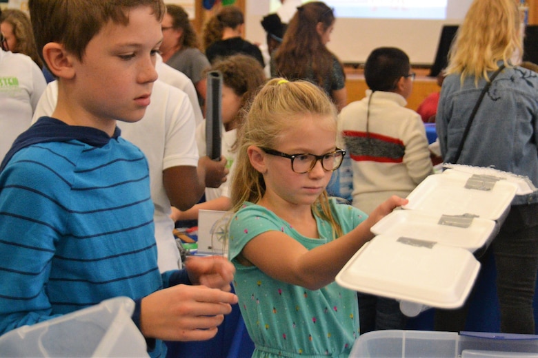 girl holds up floating bridge model made out of foam and tape while boy looks at the water the model in going into