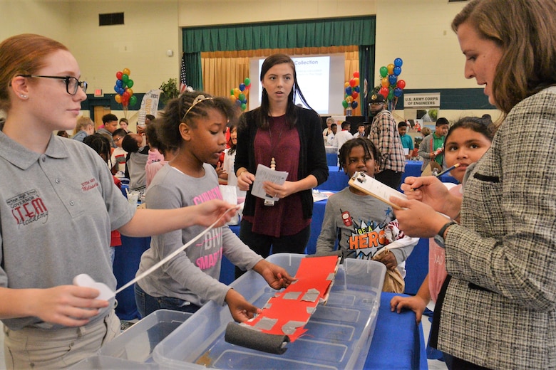 students gather around to test the model of a floating bridge in a plastic container filled with water. One girl is holding out a measuring tape and a woman is holding a clip board.
