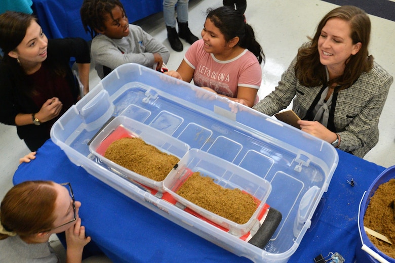 Adults and children are gathered around a container filed with water to see how much sand the model of a floating bridge can handle before it touched the bottom