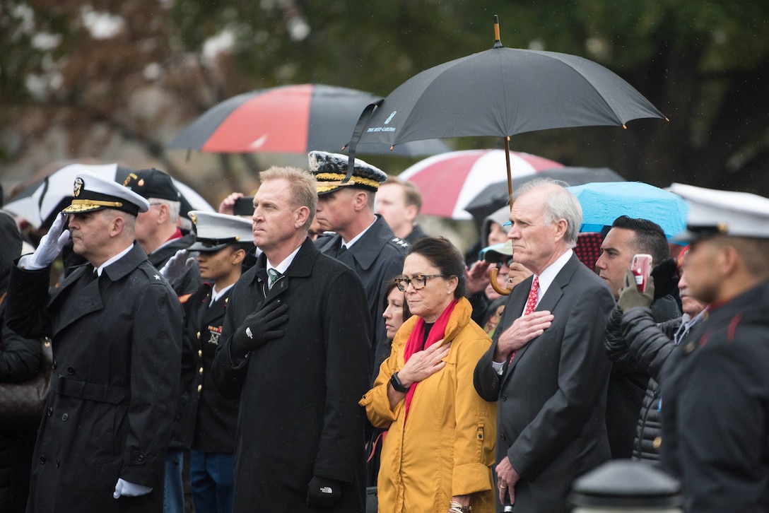 Deputy Defense Secretary Patrick M. Shanahan and others hold their hands over the hearts during a rainy outdoor ceremony.