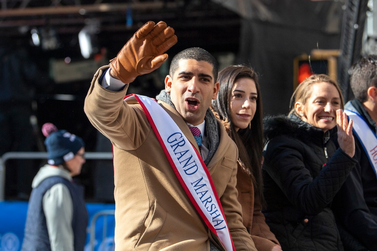 A man wearing a grand marshal sash waves to parade watchers from a car.