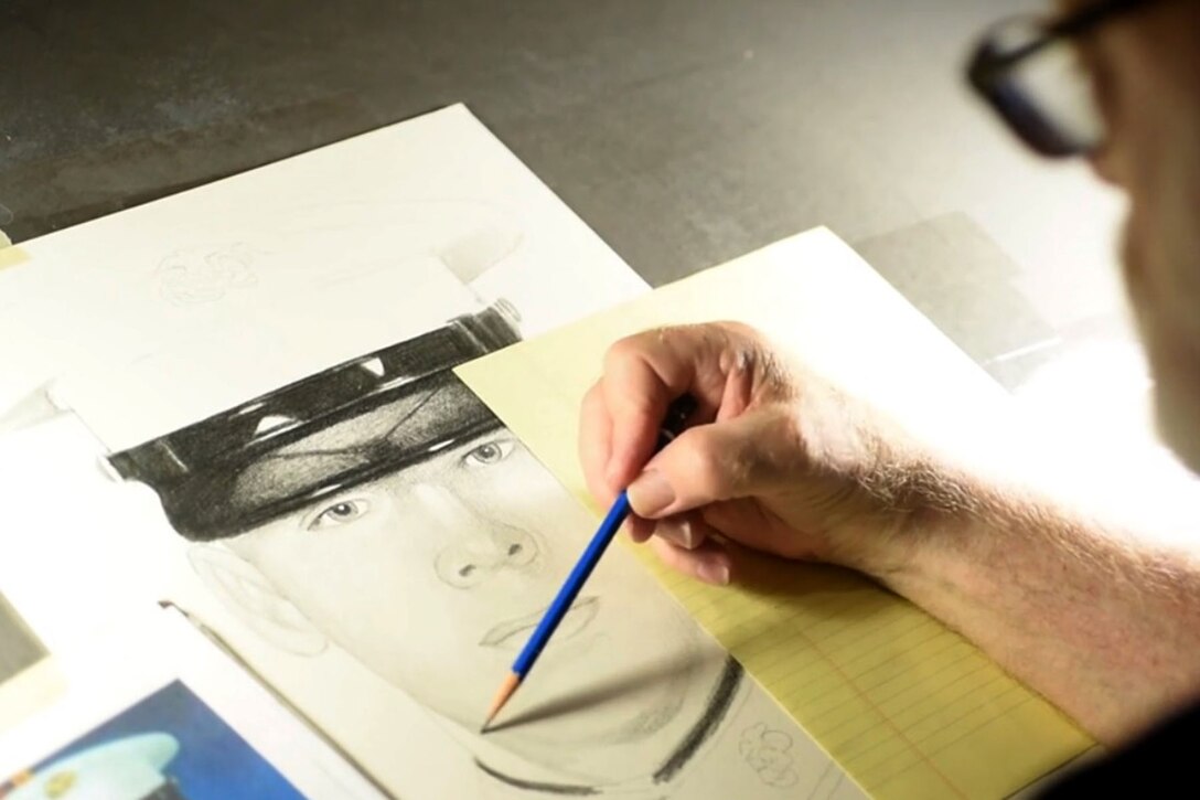 A man has a pencil to paper, drawing a portrait based on a photo of a fallen service member that sits beside the drawing pad.