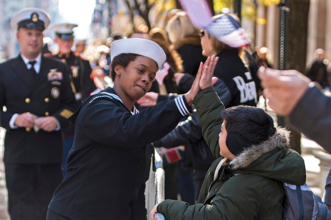 A sailor high-fives a boy along a parade route.