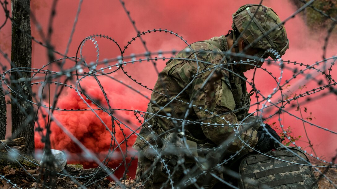 A combat medic kneels on ground working on equipment with red smoke in the background