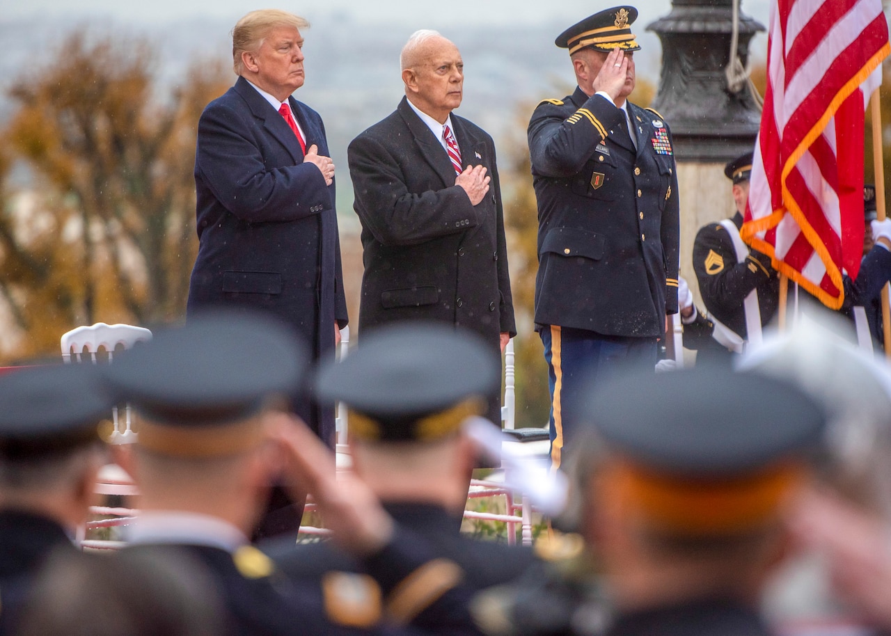 President Trump and another man stand on a dais holding their hands over their hearts while another man in uniform is saluting