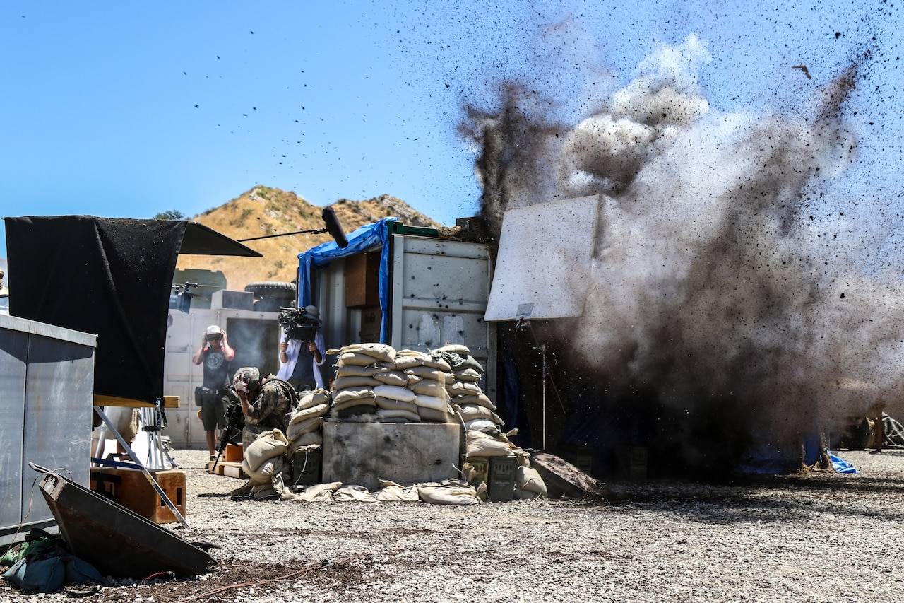 A camera crew films an actor taking cover from an explosion near a shipping container.