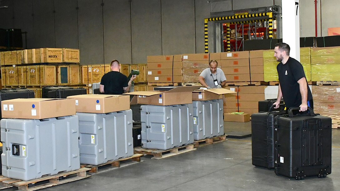 Workers in a warehouse inspect and move packing kit crates in rows.