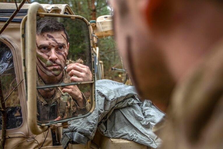 A soldier looks in a vehicle's side-view mirror while applying face paint.