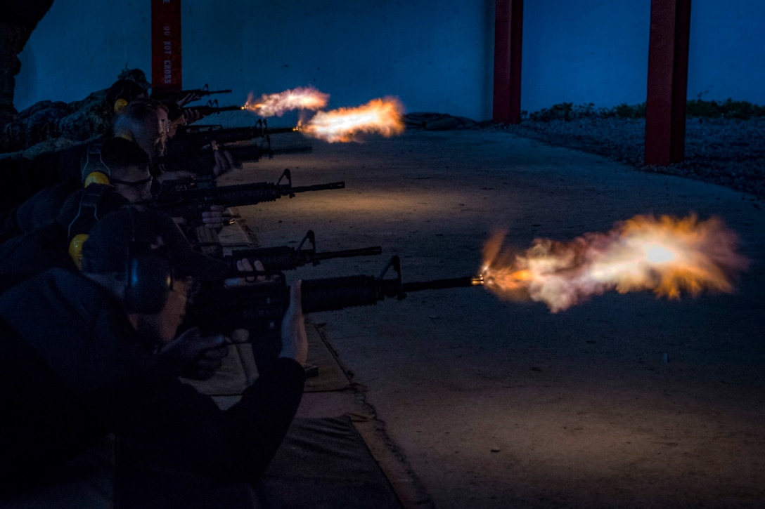 Sailors fire weapons on a firing line against a dark blue sky.