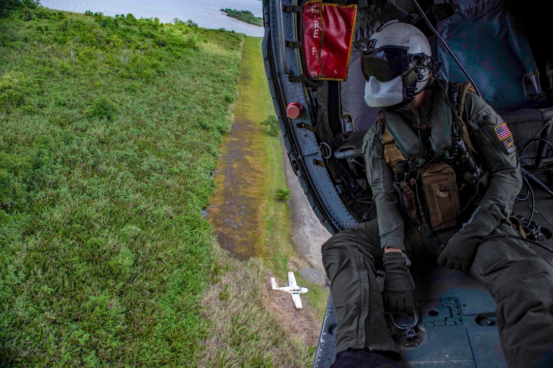 A sailor looks out from a helicopter flying over grasslands.