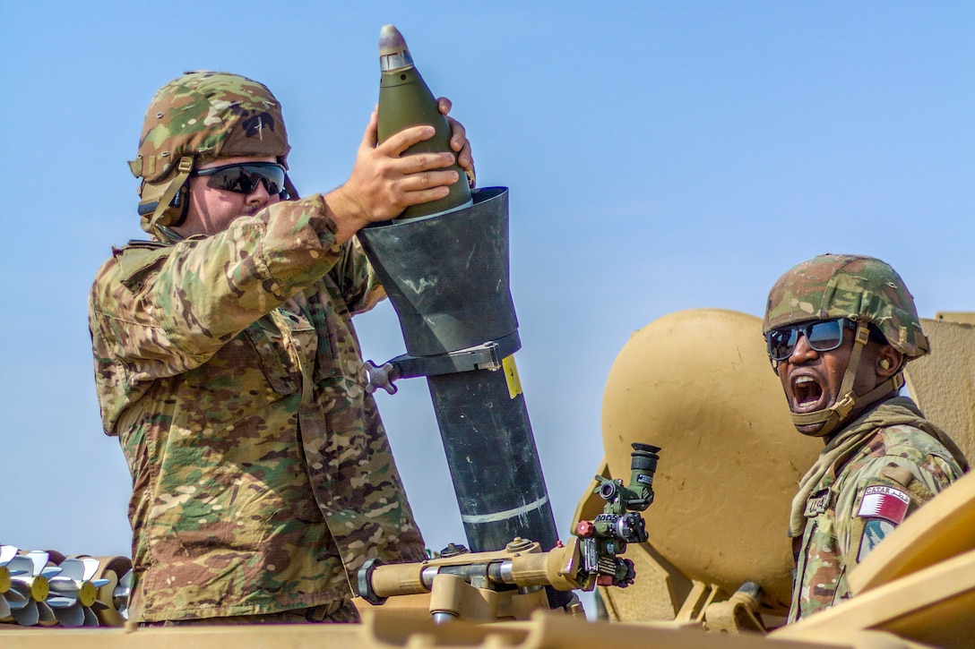 A soldier loads a mortar while another communicates with the first.