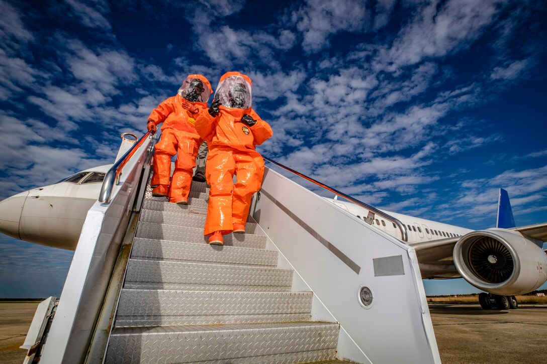 Two soldiers in protective orange bodysuits walk down the stairs of an airplane.
