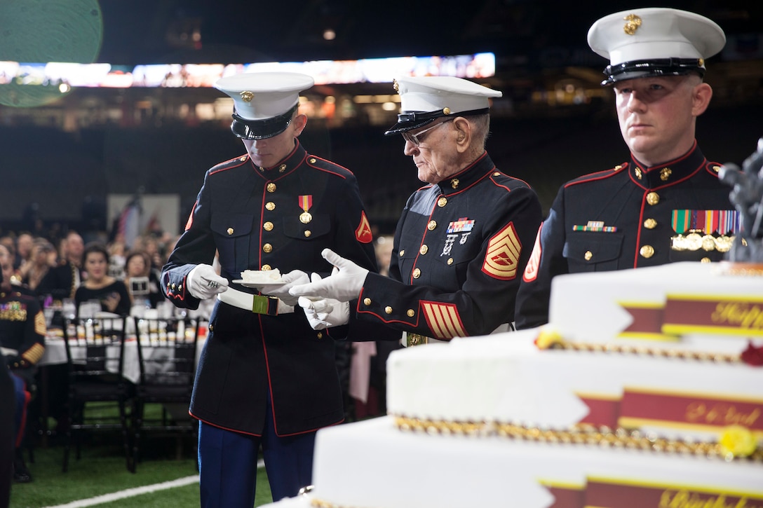 The oldest Marine present, retired Gunnery Sgt. Robert Allen, center, passes a slice of cake to the youngest Marine present, Lance Cpl. Ryan Fisher, left, an administrative specialist with G-1, 4th Marine Division, during the 243rd annual Marine Corps birthday ball at the Mercedes-Benz Superdome on Nov. 16, 2018. The passing of the cake from the oldest to the youngest Marine symbolizes the passing of history and traditions to the next generation. (U.S. Marine Corps photo by Lance Cpl. Tessa D. Watts)
