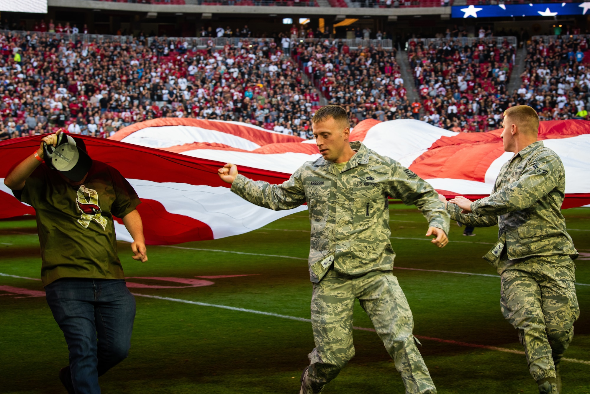 Tech. Sgt. Darren Kressin, 310th Aircraft Maintenance Unit F-16 Fighting Falcon weapons expeditor, helps unfurl an American flag during the Arizona Cardinals Salute to Service football game, Nov. 18, 2018 at the State Farm Stadium, Glendale, Ariz.
