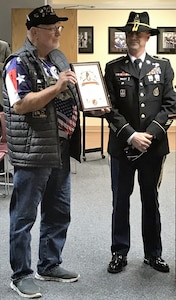 Soldier in dress uniform with a cavalry hat on presenting a certificate to a veteran with a veteran hat on.