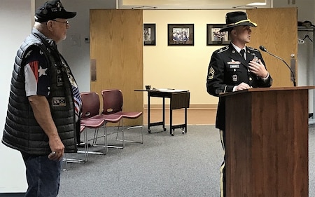 Soldier in dress uniform giving a speech in front a podium with a veteran standing behind him wearing a Vietnam veteran hat.