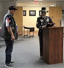 Soldier in dress uniform giving a speech in front a podium with a veteran standing behind him wearing a Vietnam veteran hat.