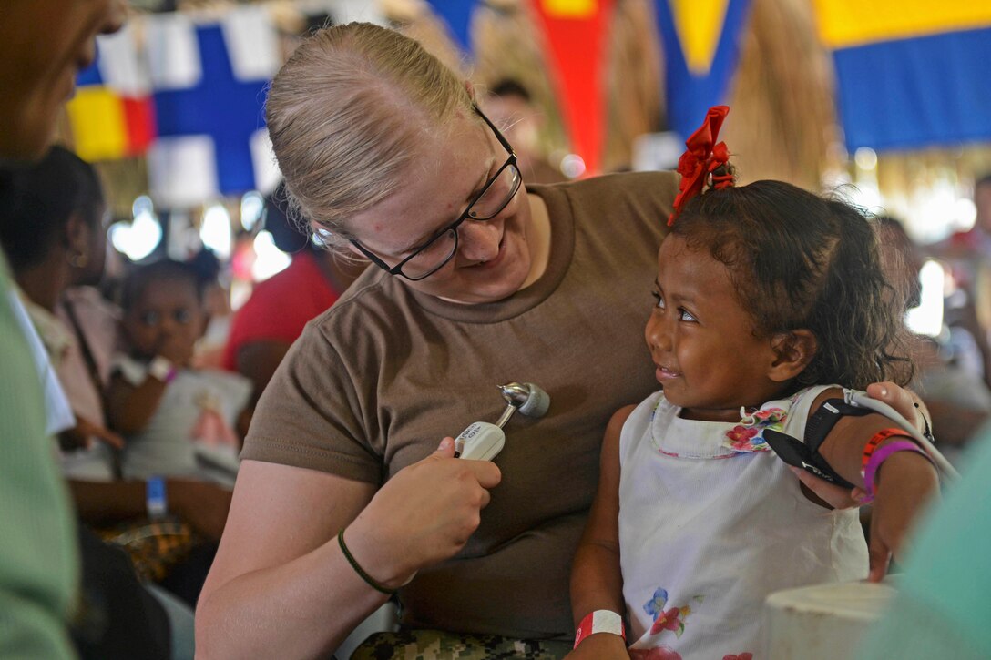 A sailor smiles at a child seated on her lap while holding a medical instrument.