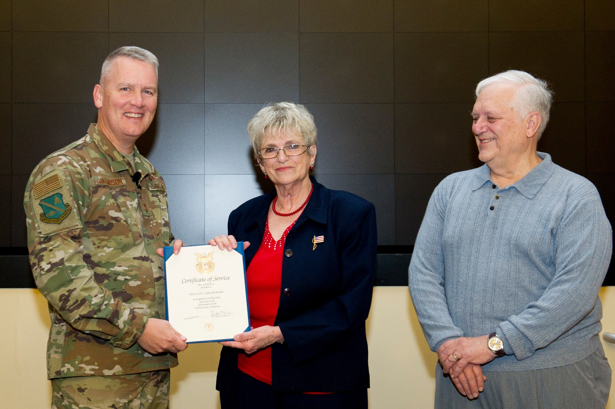 Maj. Gen. James A. Jacobson, Air Force District of Washington commander, left, presents Phyllis Grabowski, AFDW command surgeon's administrative assistant, with a 50-Year Service Certificate of Appreciation as her husband, Chester Grabowski, watches during commander's call at Joint Base Andrews, Md., Nov. 20, 2018. (U.S. Air Force photo by Master Sgt. Michael B. Keller)