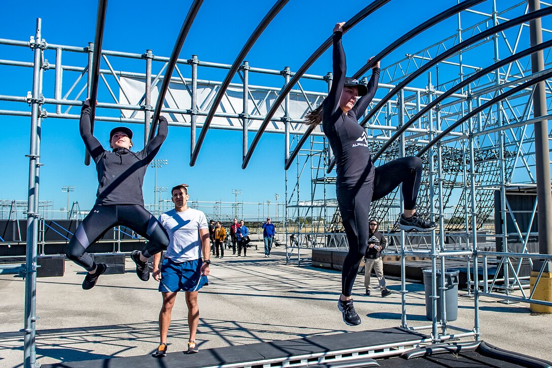 Two airmen cross an overhead parallel bar obstacle under open sky.