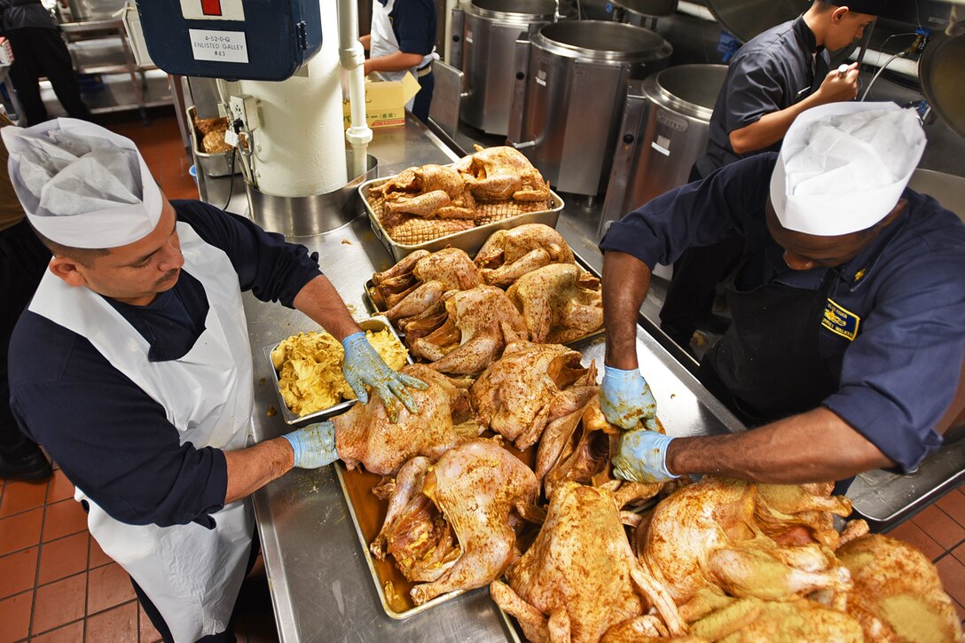 Several sailors prepare turkeys laying on a countertop on a ship.