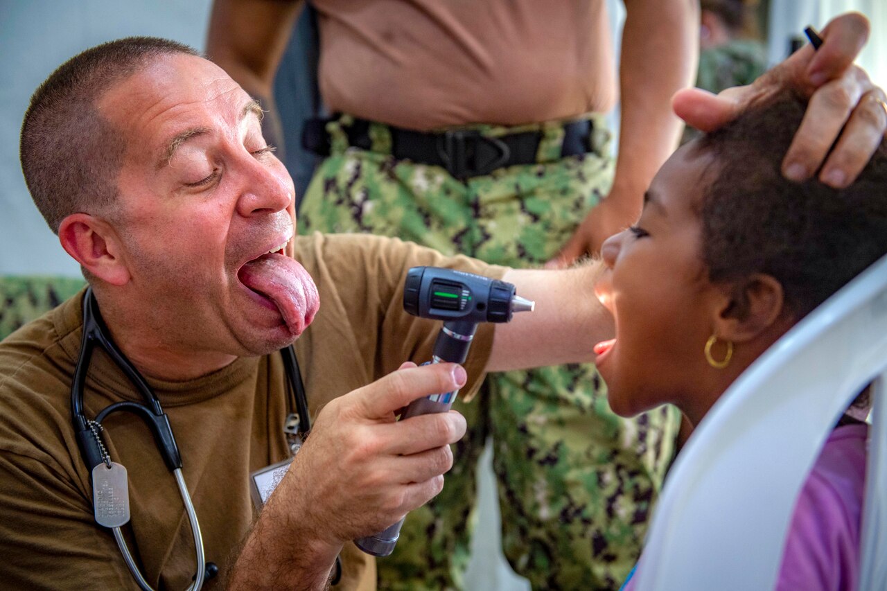 A sailor opens his mouth and sticks out his tongue to show his young patient what to do while he examines her.