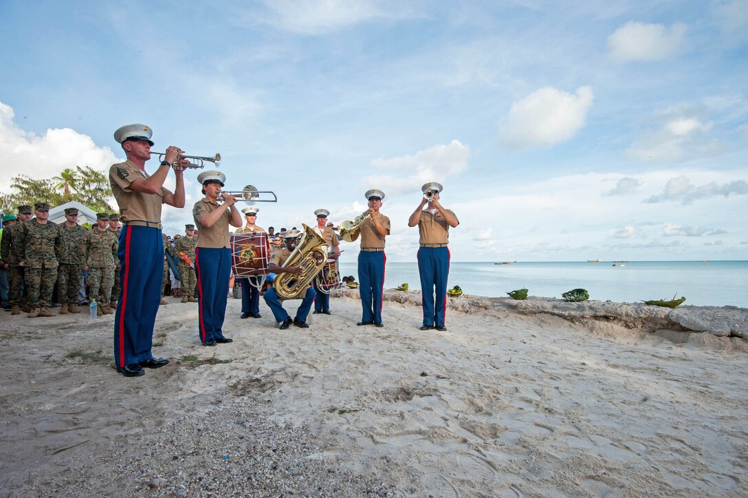 A Marine corps band perform on the beach while others watch in the background.