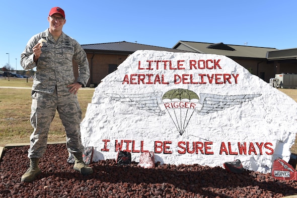 A man wearing a red hat and a U.S. Air Force Airman Battle Uniform stands to the left of a white rock that says 'Little Rock Aerial Delivery, I will be sure always.' The man holds a white plastic piece shaped like a 'U' in his right hand.