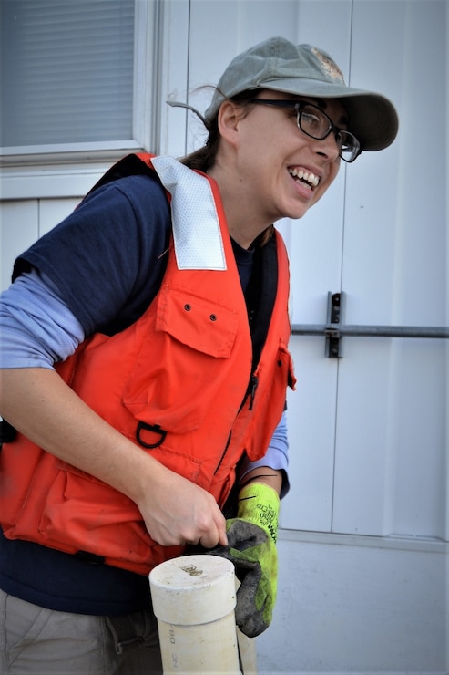 Woman in orange life vest and glasses smiles off camera to the right while working outside on a plastic tube used for oyster reef restoration