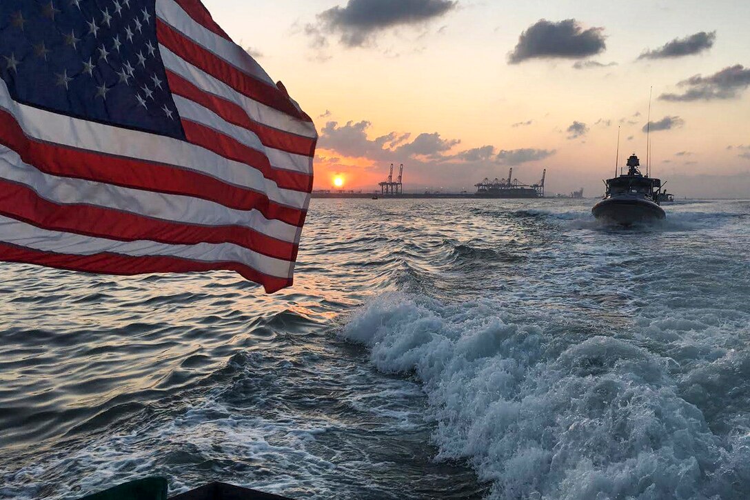 An American flag waves on a ship traveling in water as the sun sets.