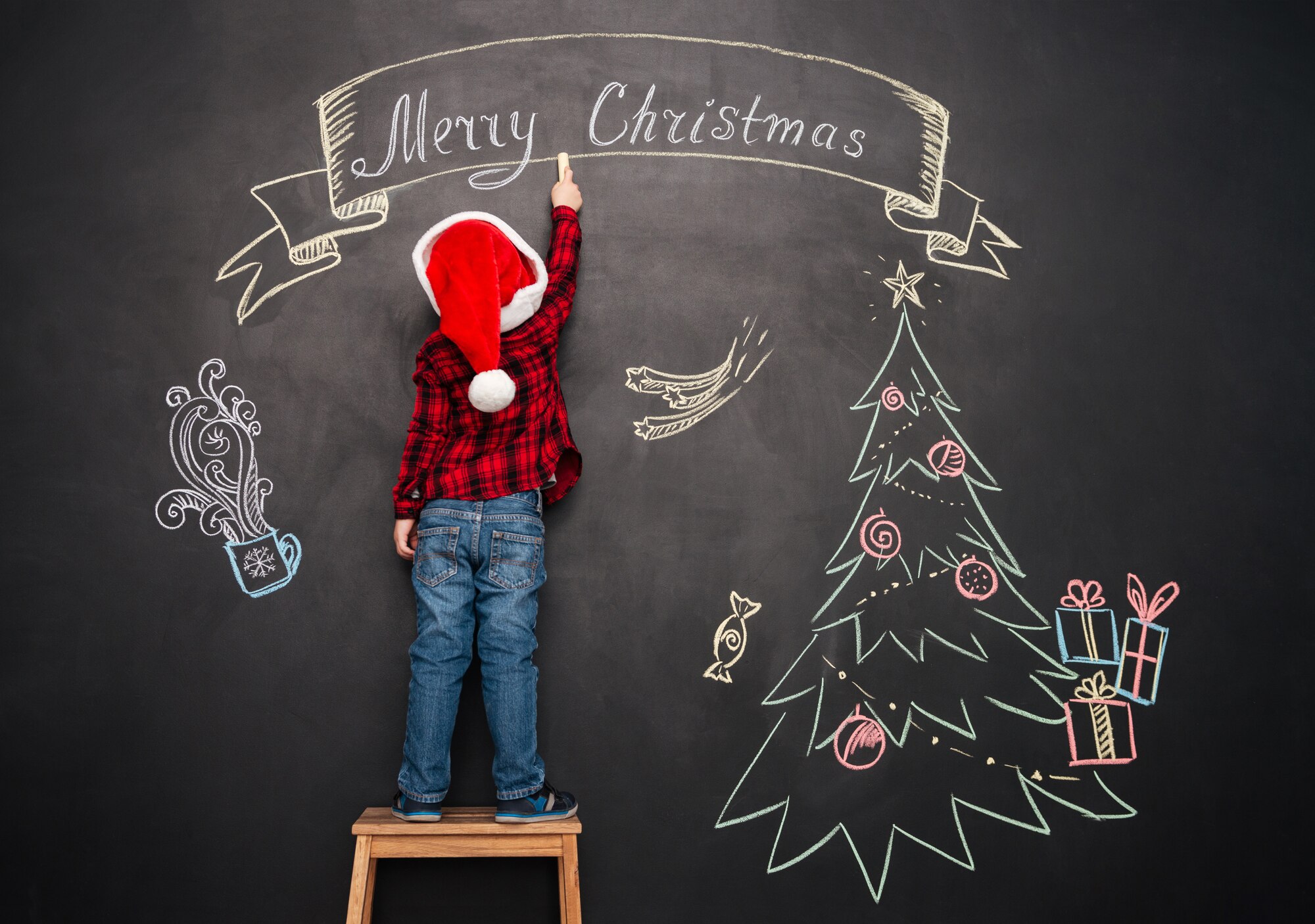 Image of little child wearing hat standing on stool near Christmas tree while drawing on blackboard.