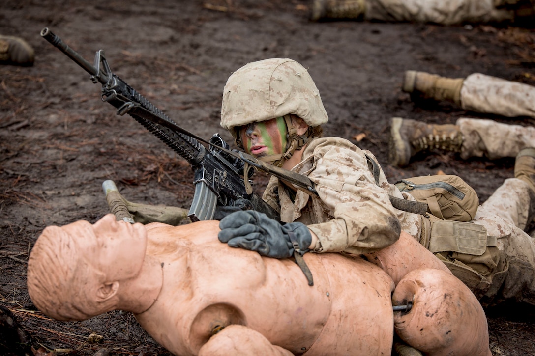 A Marine with a rifle rests her arm on a mannequin on the ground.