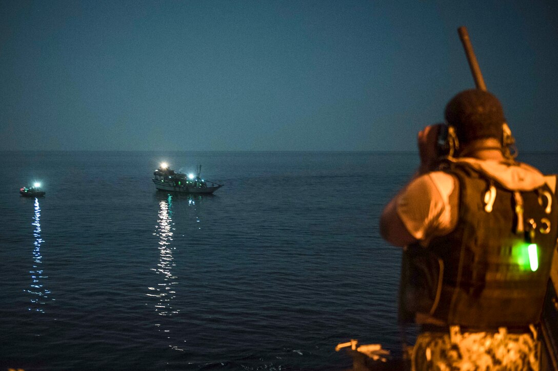 A sailor looks out at a small vessel in the sea at night.