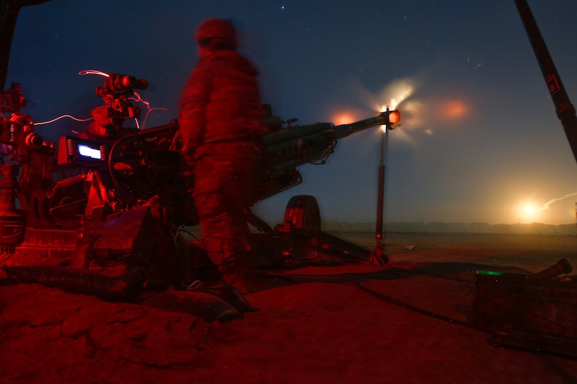 Soldiers fire a howitzer at night, illuminated by red light.