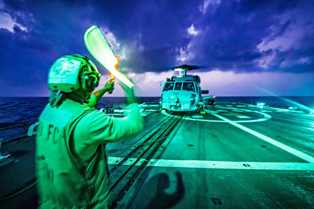 A sailor signals a helicopter on the flight deck of a ship all bathed in green light.