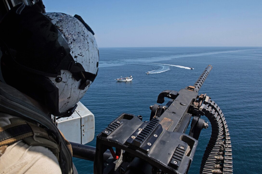 A sailor manning a machine gun looks out from a helicopter boats on the ocean below.