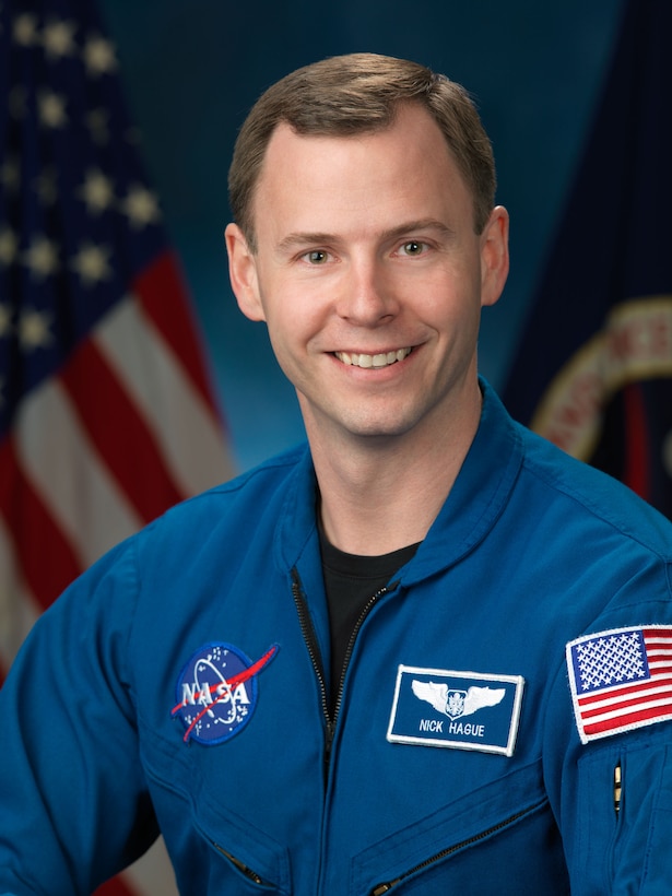 Air Force Col. Nick Hague poses in an official NASA photo. He wears a blue flight suit with a NASA logo and a name badge on the front.