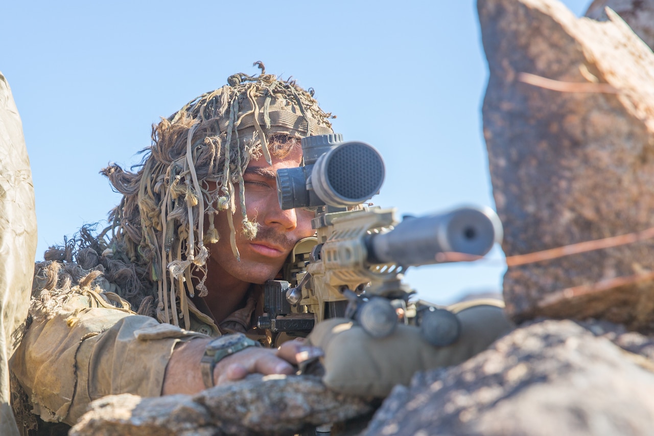 A marine takes aim at a distant target. Wearing a rope-like camouflage, he closes one eye and peers through his rifle scope.