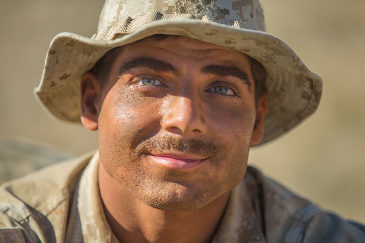 Marine Lance Cpl. Logan Stutte poses for the camera wearing a floppy desert-colored camouflage hat. His face is covered in stubble and dirt.