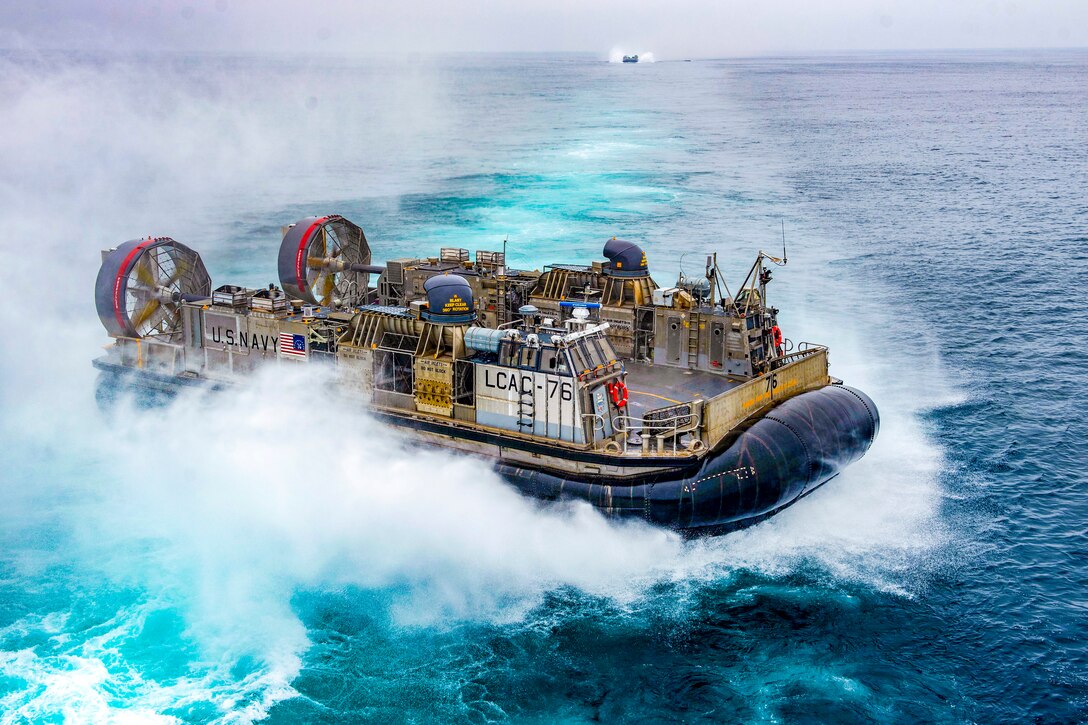 A landing craft blows clouds of water up as it approaches a larger naval vessel.