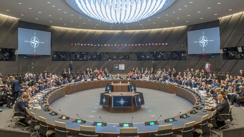 Defense ministers meet at the NATO headquarters in Brussels. A large conference room features a round table in the center of the image. More than 100 people are present.
