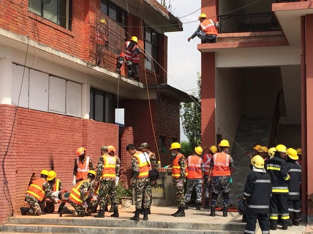 First responders in orange vests and camo pants gahter outside a building, as several of them maneuver on second-floor structures.
