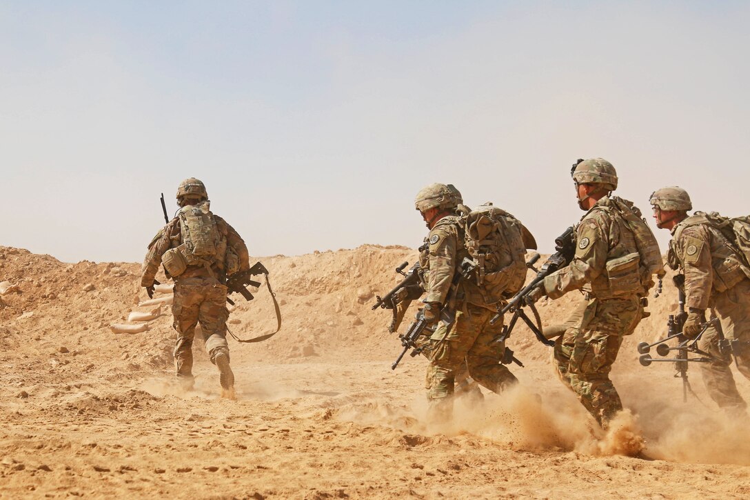 A group pf soldiers race across dusty soil while holding equipment.