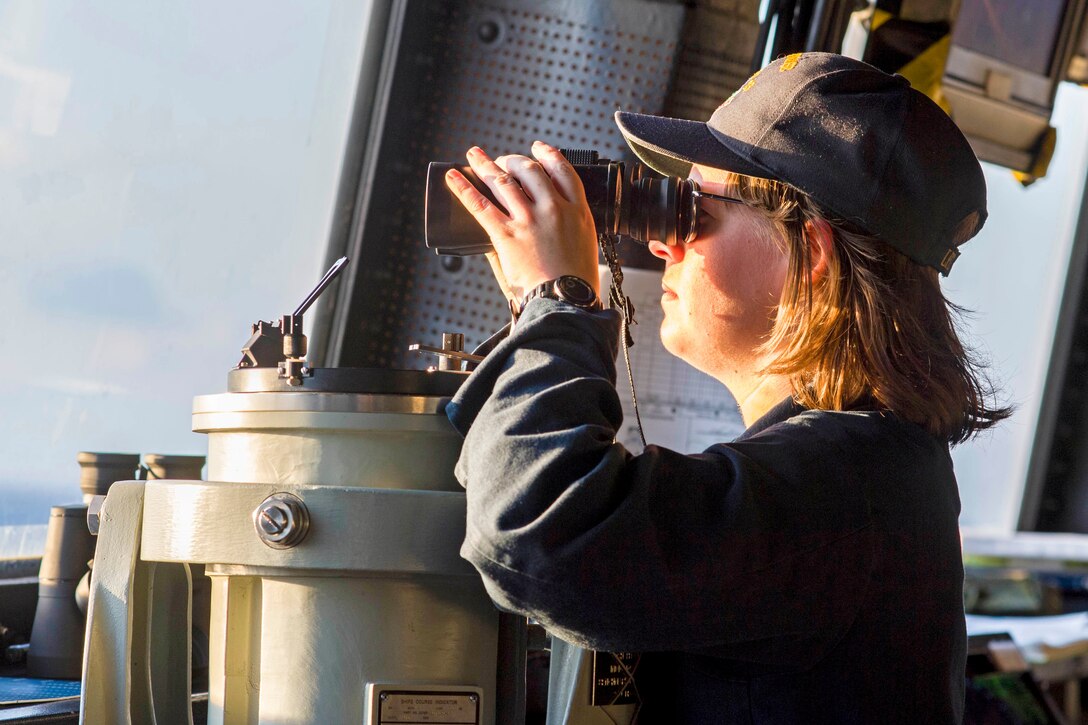 A sailor stands watch using binoculars.