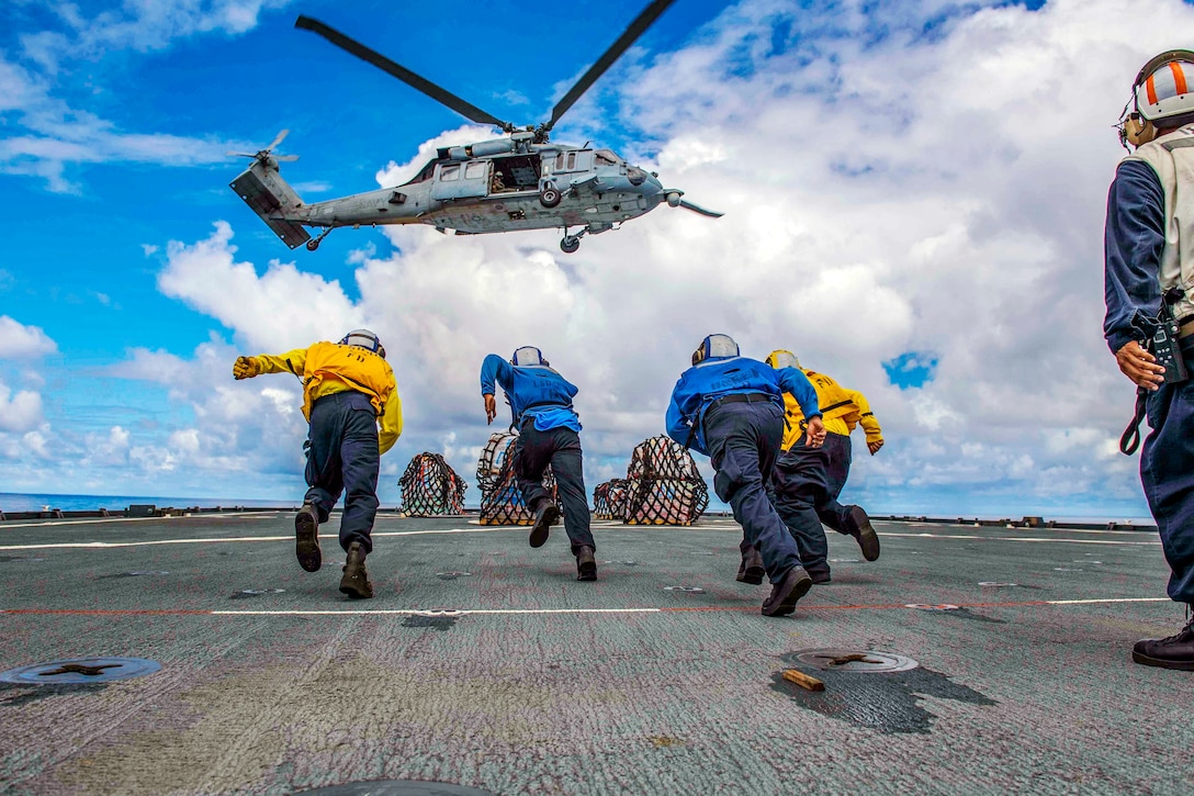 Sailors run on a ship's flight deck toward a hovering helicopter.