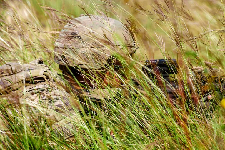 A soldier crouches in tall grass while aiming his gun looking through his sights.
