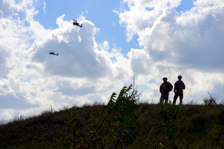 Soldiers stand on a hilltop as helicopters fly overhead.
