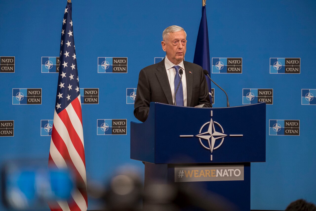 Defense Secretary James N. Mattis speaks at NATO. Behind him is a blue backdrop with the NATO logo. On either side of him are the U.S. flag and the NATO flag.