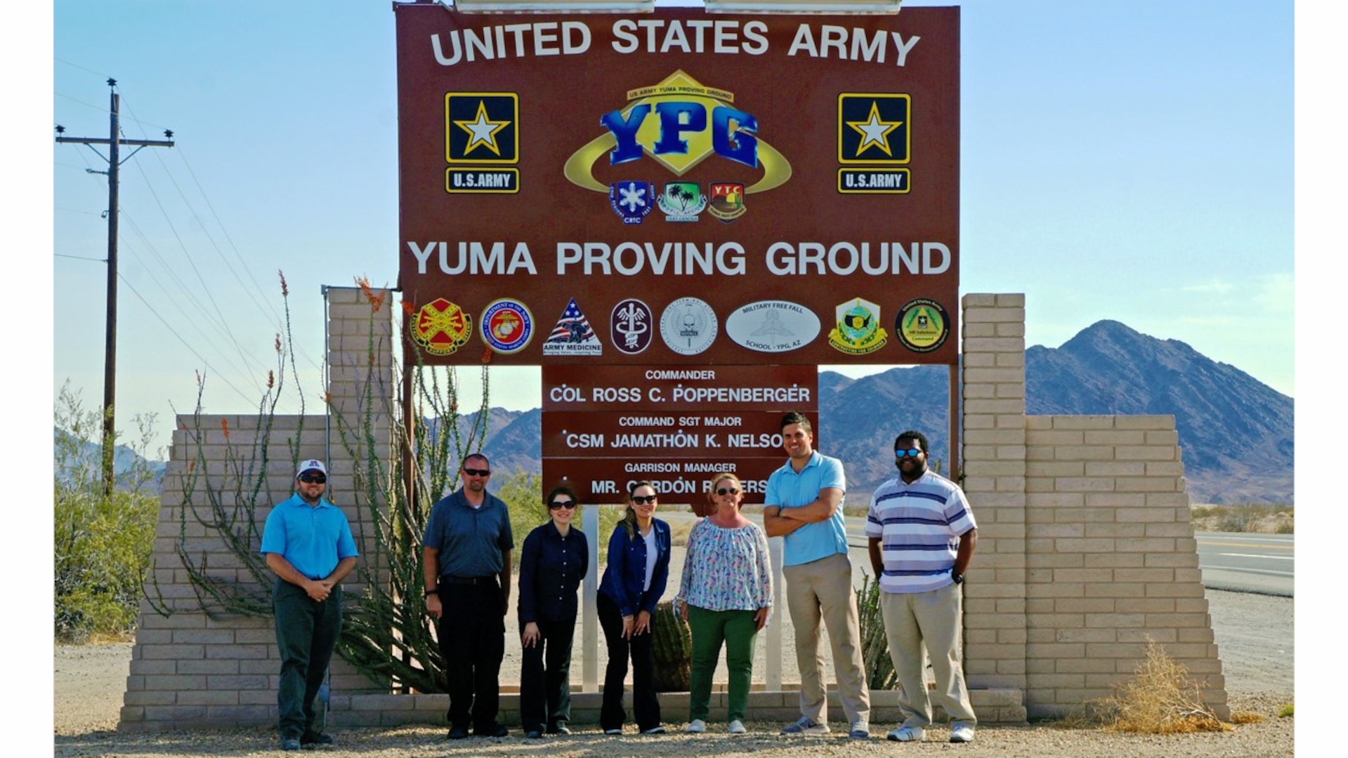 Seven people stand in front of a large sign at the entrance to Yuma Proving Ground.